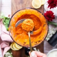 A spoon showing close up the texture of the romesco sauce when processed shwoing the vibrant orange color and how slightly chunky it is