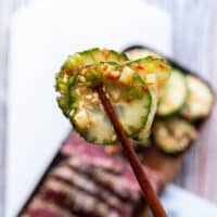 a hand holding a cucumber using chopsticks up close showing the texture and marinade of the cucumber salad