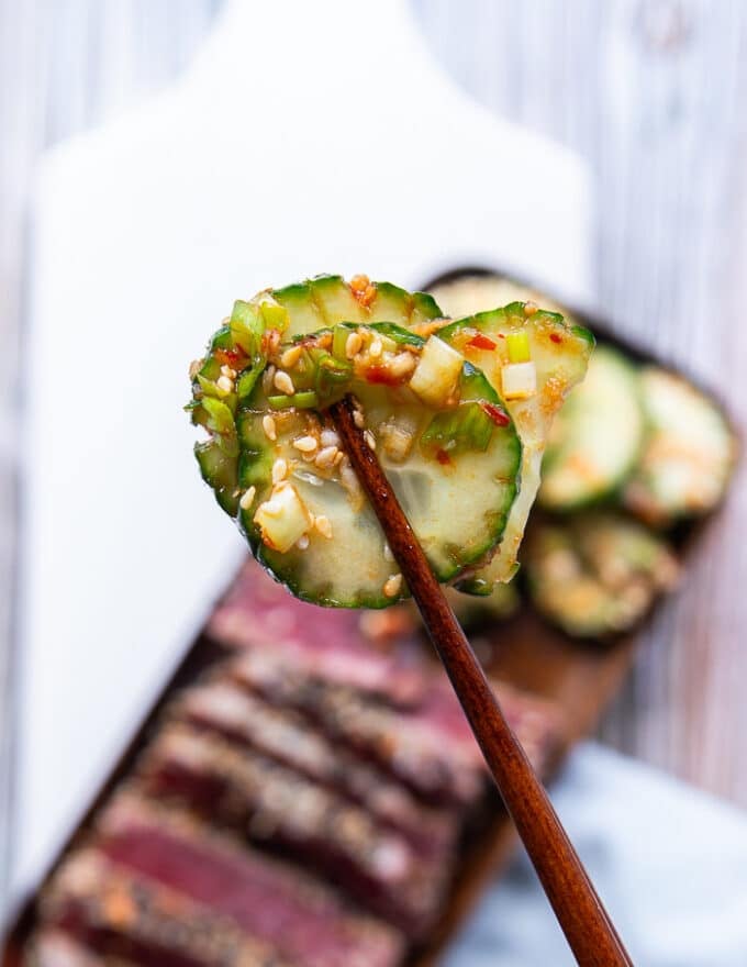 a hand holding a cucumber using chopsticks up close showing the texture and marinade of the cucumber salad
