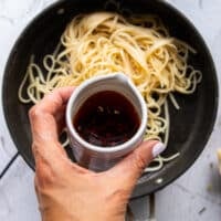 A hand pouring the spicy chili oil over cooked pasta to make chili crab pasta