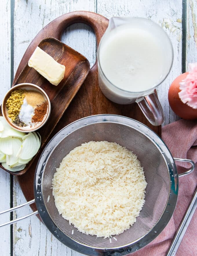 Ingredients to make coconut rice on a cutting board including rinsed long grain rice in strainer, some butter in a bowl, a bowl of spices, some sliced onions and a jar of coconut milk