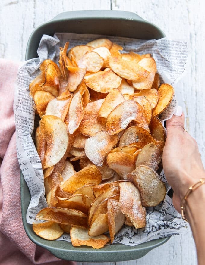 A hand holding a deep plate of homemade potato chips with a paper lining 