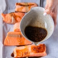 A hand pouring half of the maple glaze form the bowl on to the salmon fillets on a baking sheet