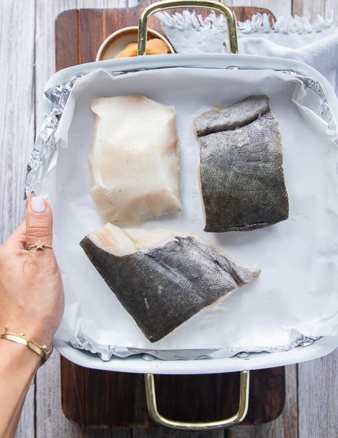 A hand holding a tray of black cod showing what the fish is and how black the skin is and how thick the fillet is. 