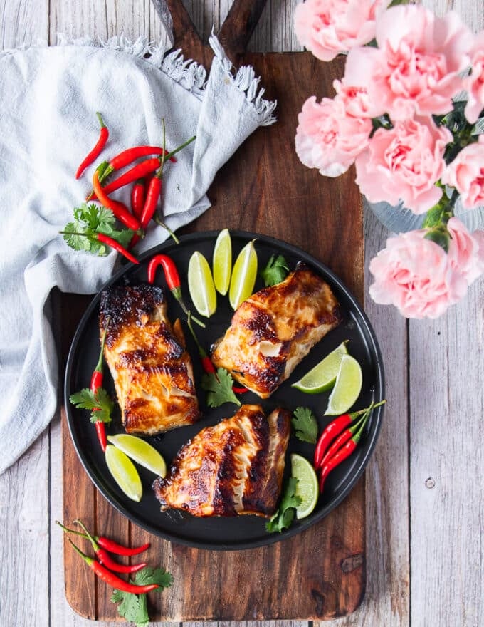 A full plate of miso black cod with lime wedges and a tea towel on a kitchen table and some flowers on a vase 