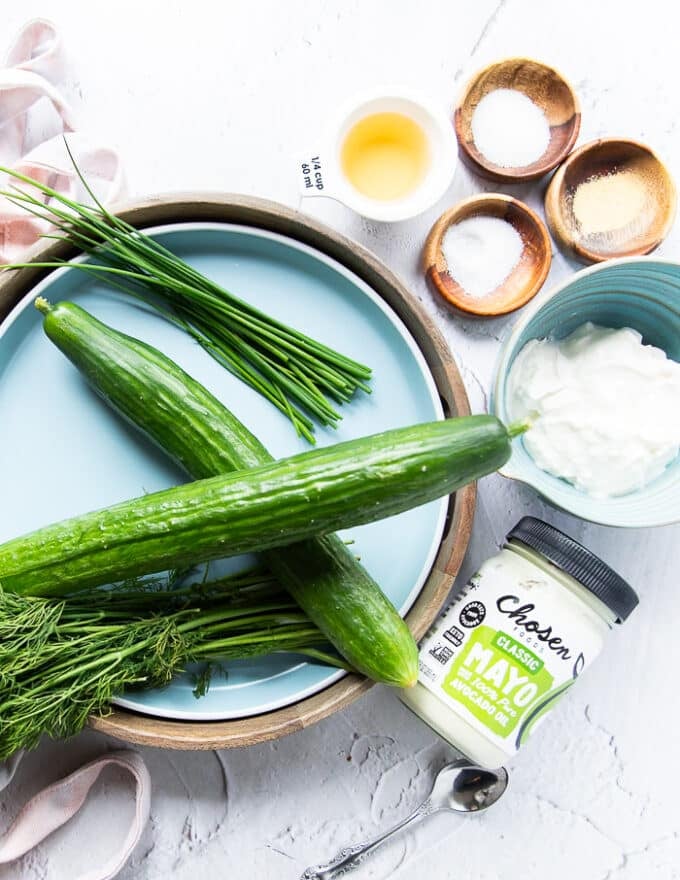 Ingredients for creamy cucumber salad including two large cucumebrs, a bowl of sour cream a few tablespoons mayo, some garlic powder and seasoning, lemon juice, a bunch of fresh dill