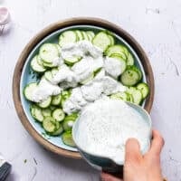 A hand pouring the creamy dressing over the cucumber, dill and onions