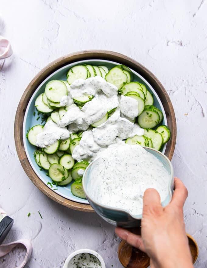 A hand pouring the creamy dressing over the cucumber, dill and onions