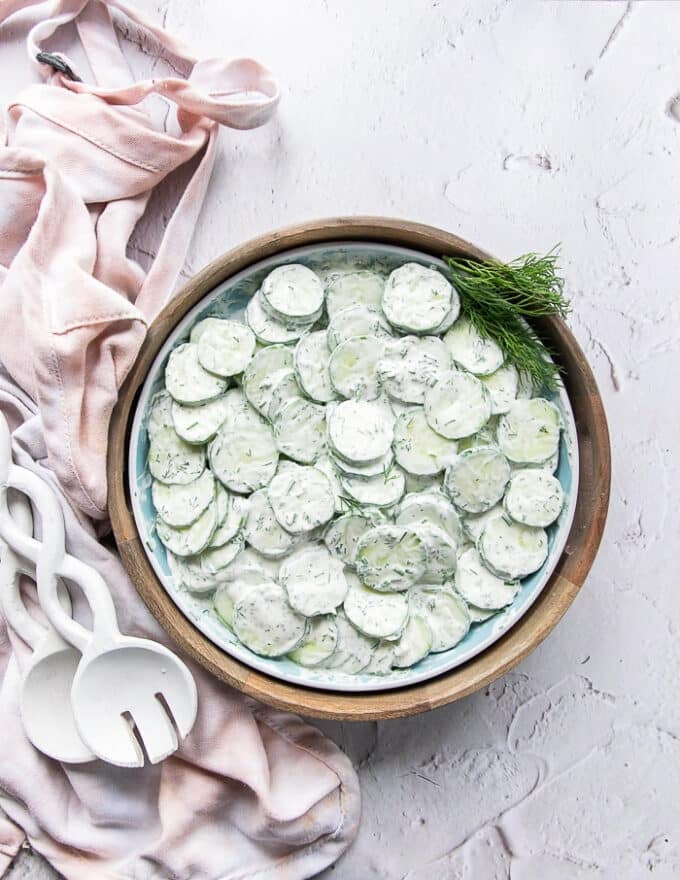 Top view of final creamy cucumber salad surrounded by salad spoons and a kitchen towel garnished with lots of fresh dill