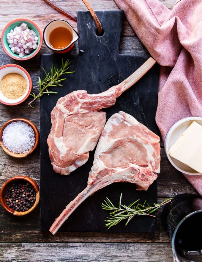 Ingredients for grilled veal chops on a board including seasoning in small bowls, some balsamic vinegar, some shallots minced, butter in a bowl, fresh rosemary sprigs, honey in a small bowl and two veal chops
