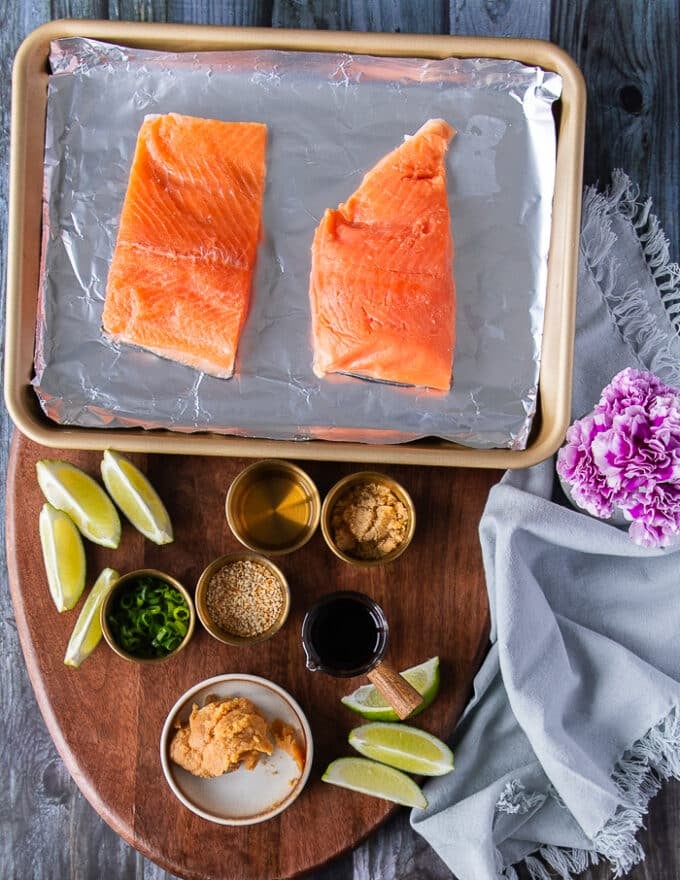 Ingredients needed for miso glazed salmon including the salmon fillets, some miso paste in a bowl, soy sauce in a bowl, some sake or mirin in a bowl, sesame seeds in a bowl and some lime for garnish