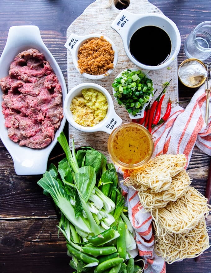Ingredients to make Mongolian Ground Beef Noodles on a board including a bowl of ground beef, a bowl of soy sauce, a cup of stock, a bowl of brown sugar, a cup of water and table spoon of cornstarch , a bowl of green onions, a bowl of minced garlic and ginger, some noodles cooked and some chillies