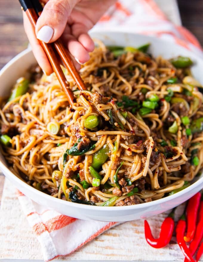 A hand holding two chopsticks stretching the Mongolian Ground Beef Noodles on a plate showing the ground beef, the green onions and chilli flakes over the noodles