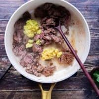 The ground beef is added to a skillet with ground ginger and garlic and a wooden spoon is mixing them