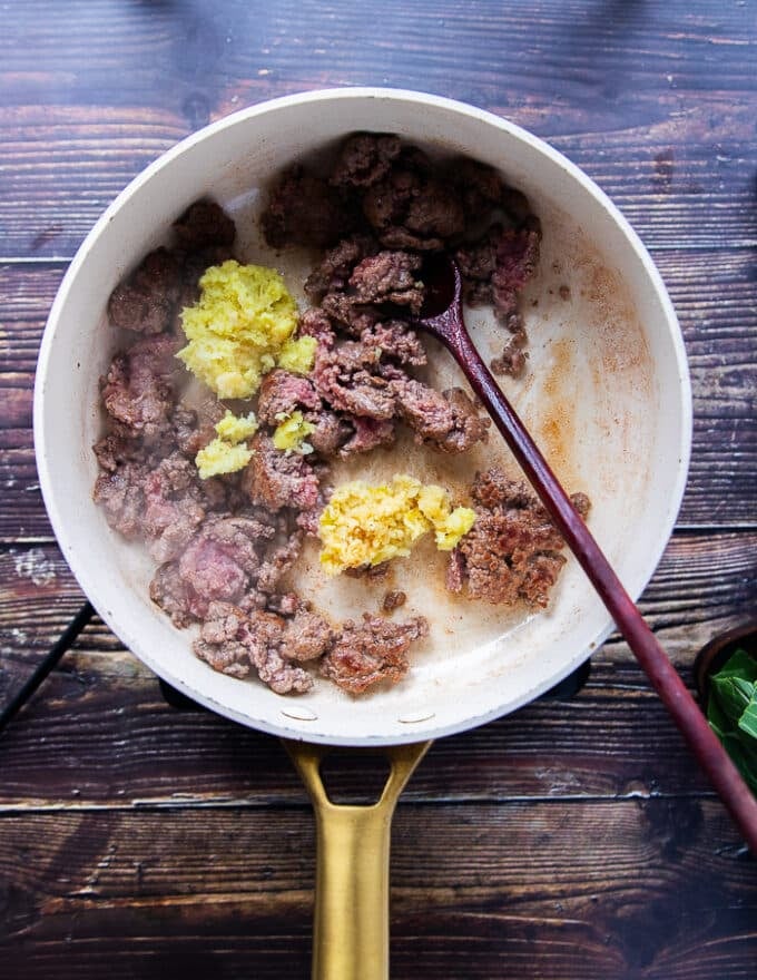 The ground beef is added to a skillet with ground ginger and garlic and a wooden spoon is mixing them