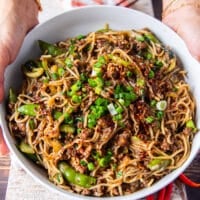 two hands holding a bowl of Mongolian Ground Beef Noodles garnished with lots of green onions and chilli flakes
