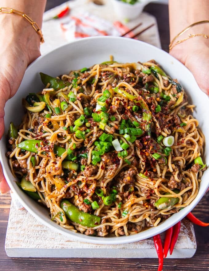 two hands holding a bowl of Mongolian Ground Beef Noodles garnished with lots of green onions and chilli flakes