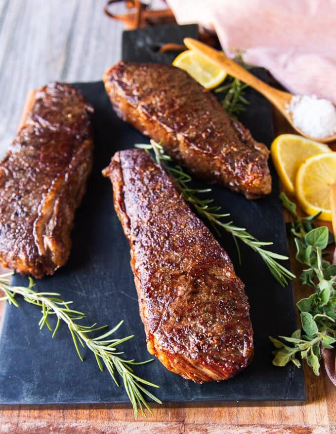 Three steaks on a board revere seared and perfectly browned on the outside and surrounded by lemon and rosemary 