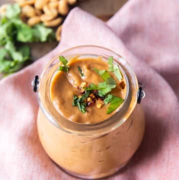 A jar of satay sauce garnished with crushed peanuts, cilantro and chilli flakes
