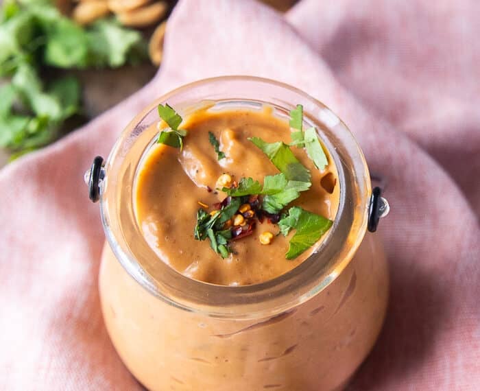 A jar of satay sauce garnished with crushed peanuts, cilantro and chilli flakes