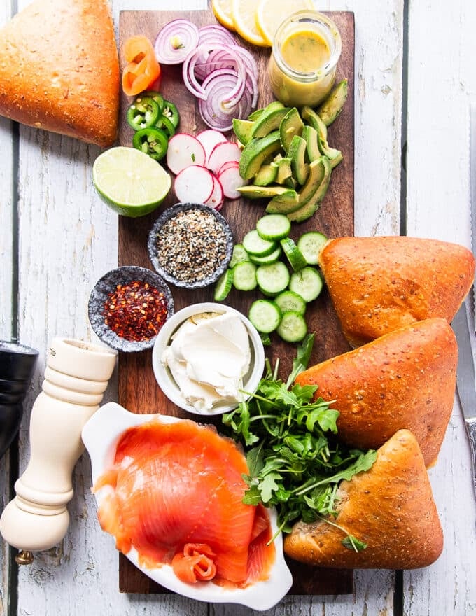 Ingredients for making a smoked salmon sandwich including smoked salmon on a board, some bread, some sliced cucumbers some arugula, some avocado slices, some radishes and red onion sliced and a bowl of cream cheese, a bowl of bagel seasoning and chilli flakes