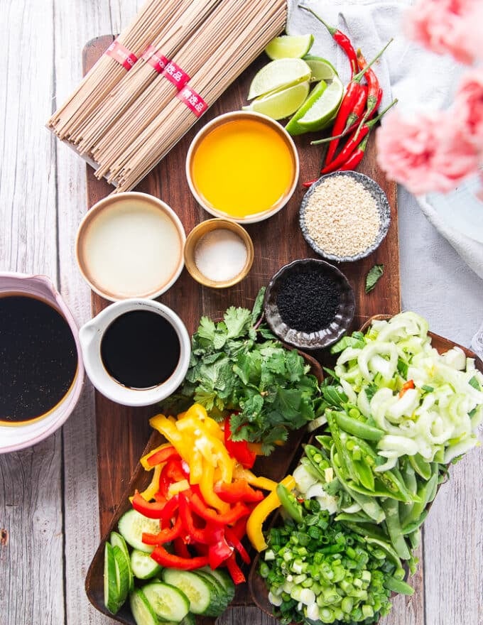 Ingredients for two soba noodles recipes including a bowl of soy saucem a bowl of brown sugar, some rice wine vinegar, some miso in a bowl, a plate of shredded veggies, cilantro, sesame oil, seasoning, green onions minced, toasted sesame seeds 