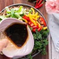 A hand drizzling the dressing over the veggies and cold soba noodles
