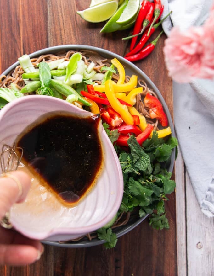 A hand drizzling the dressing over the veggies and cold soba noodles 