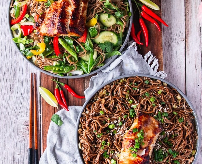 two plates of soba noodles, one served warm and the other cold with veggies