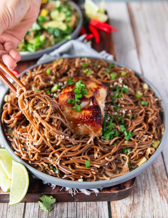 A hand holding chopsticks and stretching the soba noodles recipe showing how perfectly cooked it is with a warm sauce 