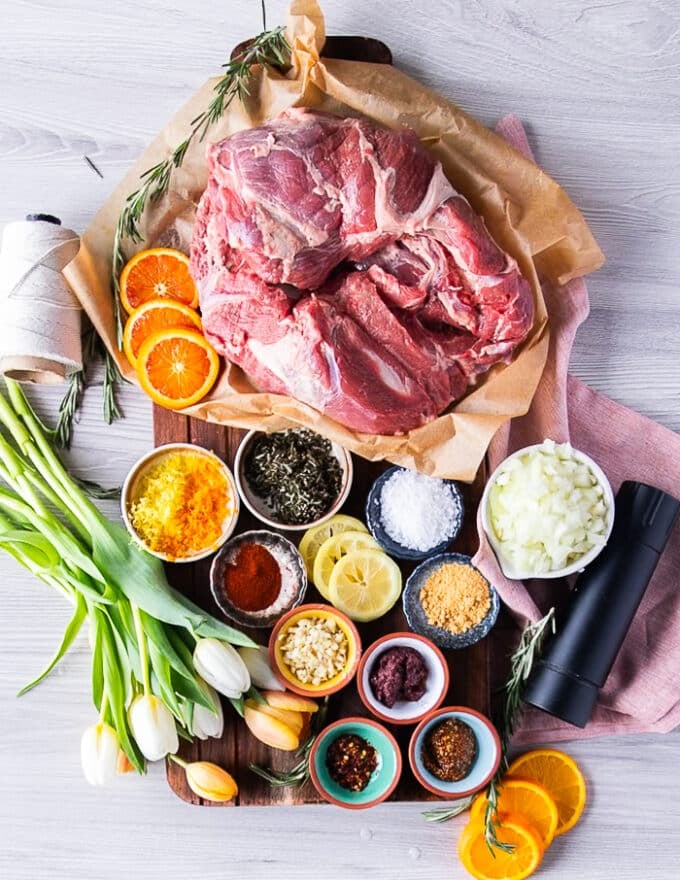 All ingredients needed for roast boneless leg of lamb including boneless leg of lamb, some citrus zests, some onions, mustard, spices in separate bowls, some fresh rosemary, some garlic, lots of olive oil. 