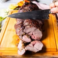 A hand holding a knife and slowly slicing the perfect boneless leg of lamb showing the first slice how perfectly cooked and flavored it looks