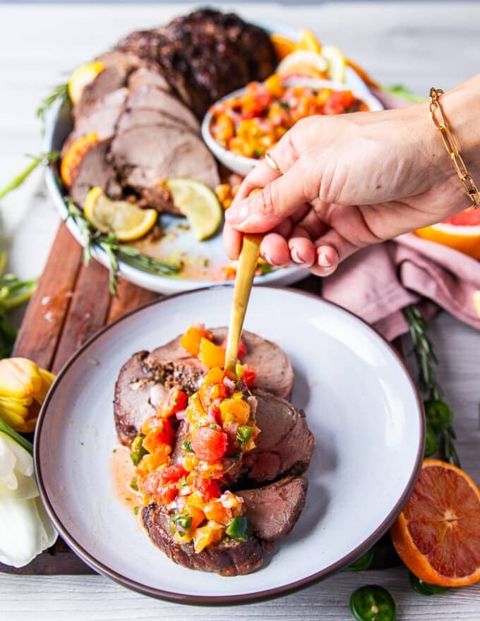 A hand pouring some citrus salsa over slices of roasted boneless leg of lamb