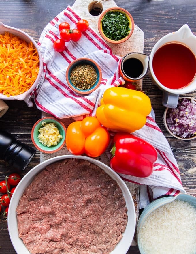ingredients to make stuffed peppers casserole including a variety of colored peppers, some ground beef, some tomatoes in a cup, some onions, grated cheese in a bowl, minced garlic, some seasoning in a bowl, some cooked rice in a bowl, diced onions,