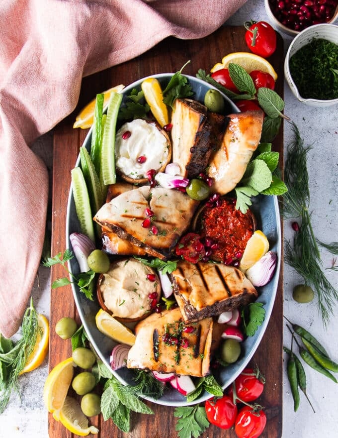 A plate of cooked arayes served with hummus bowl, yogurt bowl, sliced cucumbers, baby tomatoes and lemon wedges