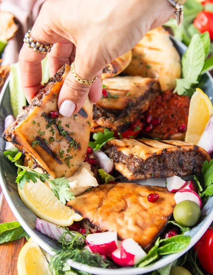 A hand holding a piece of cooked arayes and dipping it into the hummus 