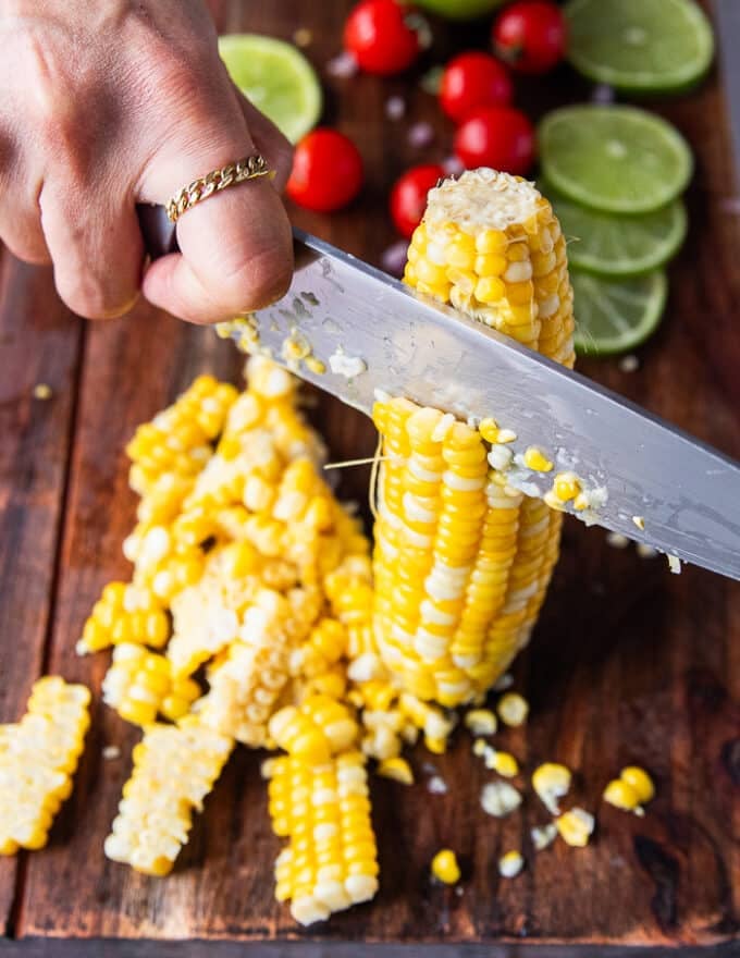 A hand holding a corn on the cob that;s cooked and a knife cutting through the kernels 