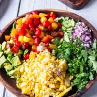 Corn and tomato salad ingredients chopped and added separately to a large bowl. Including chopped grape tomatoes, cooked corn on the cob, fresh cilantro, chopped onions, chopped cucumber and diced avocados