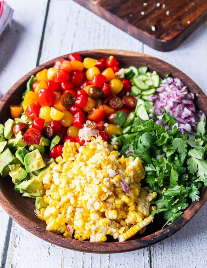 Corn and tomato salad ingredients chopped and added separately to a large bowl. Including chopped grape tomatoes, cooked corn on the cob, fresh cilantro, chopped onions, chopped cucumber and diced avocados