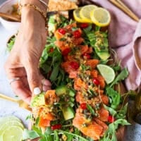 A hand holding a piece of salmon carpaccio with pita chips showing closeup of the salmon, the dressing and the capers