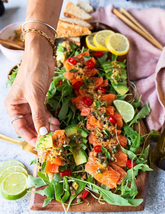 A hand holding a piece of salmon carpaccio with pita chips showing closeup of the salmon, the dressing and the capers