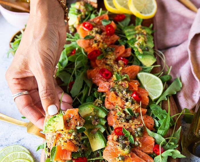 A hand holding a piece of salmon carpaccio with pita chips showing closeup of the salmon, the dressing and the capers