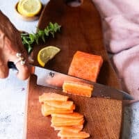 A hand holding a sharp knife and slicing the salmon against the grain into paper thin layers