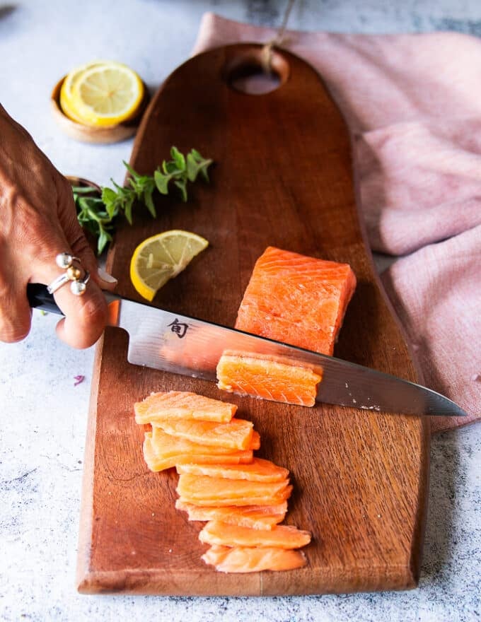A hand holding a sharp knife and slicing the salmon against the grain into paper thin layers 