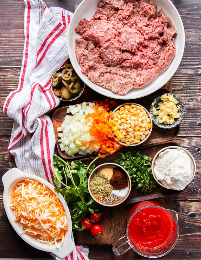 Ingredients for the taco spaghetti recipe including ground beef, some diced onions in a bowl, some minced garlic in a bowl, some diced tomatoes, some corn, some shredded cheese in a bowl, a bowl of taco spice blend, some diced peppers and oil,