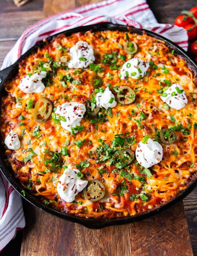 A skillet of taco spaghetti out of the oven with toppings on a wooden table and a tea towel surrounding it 