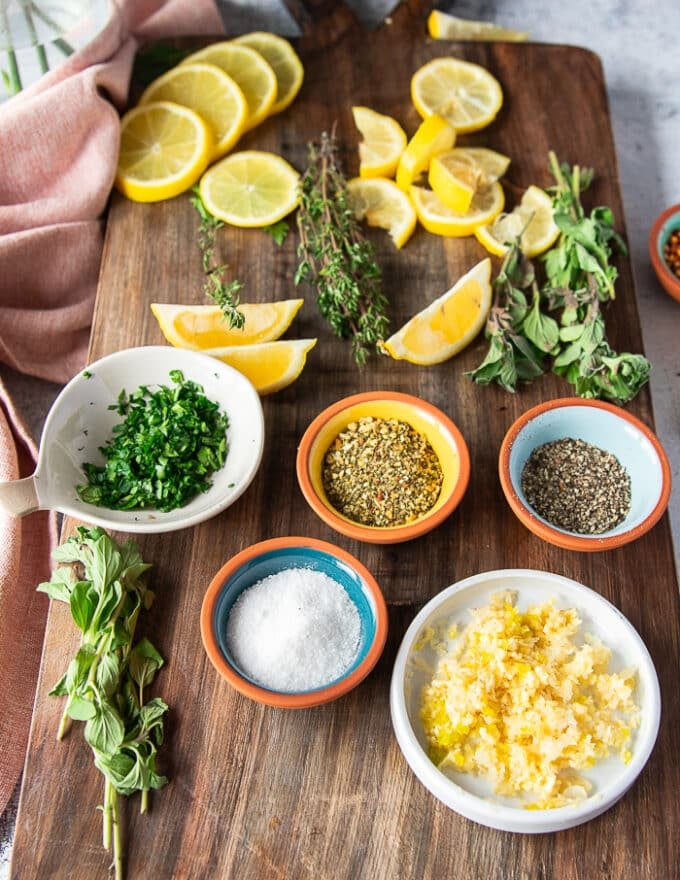 Remaining ingredients for broiled scallops in a wooden board including a dab of butter in a bowl, some seasoning in a small bowl some fresh parsley, some minced garlic on a bowl 