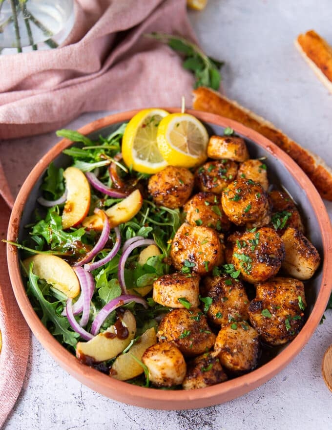 A plate of broiled scallops served with lemon slices and a side of salads