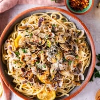 A bowl of clam pasta in a clay bowl surrounded by small dishes of herbs, lemon slices and a bowl of chilli flakes.