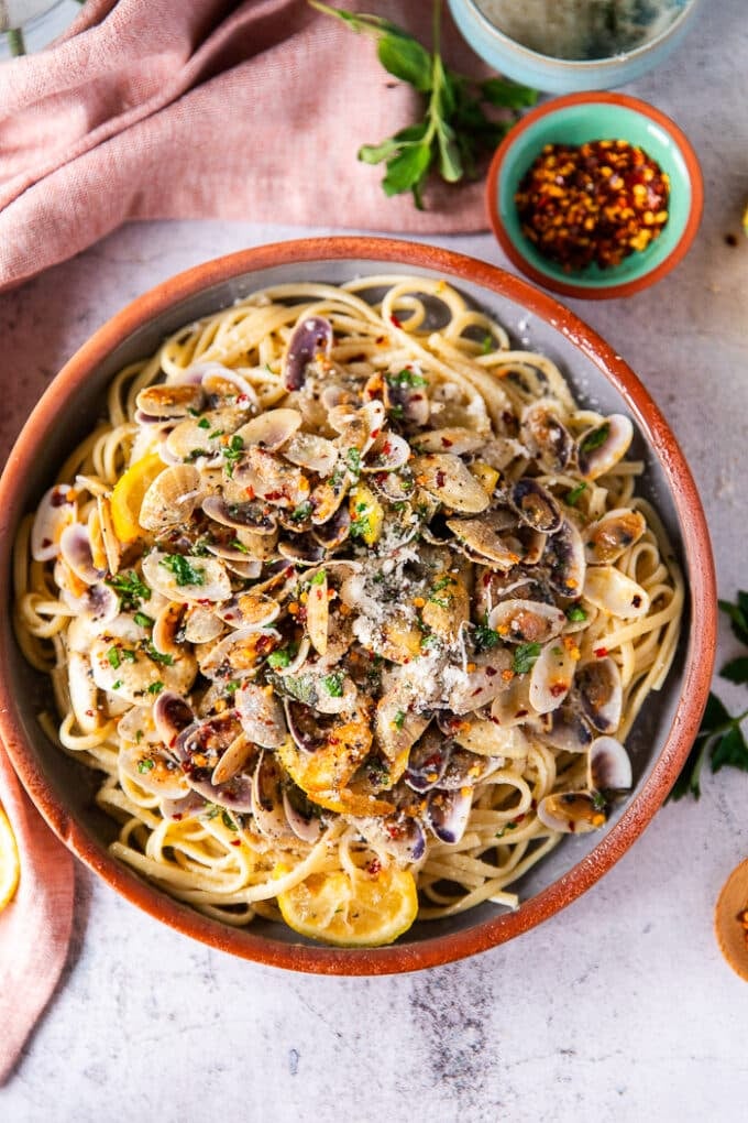 Un tazón de pasta de almeja en un tazón de arcilla rodeado de pequeños platos de hierbas, rodajas de limón y un tazón de escamas de chile. 
